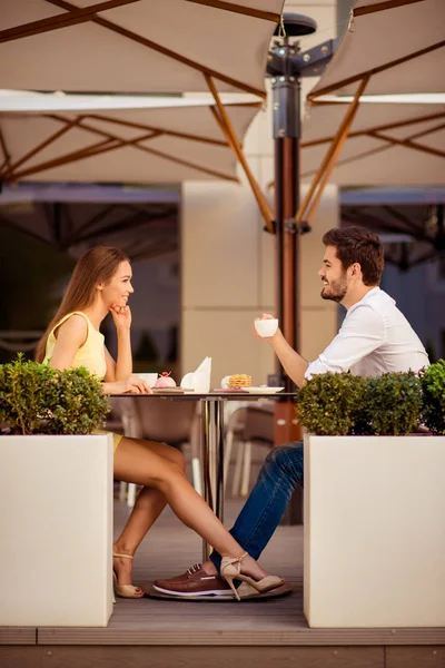 Young happy gorgeous lady is sitting in light cafe`s terrace outdoors on a sunny day and flirt with her leg with partner, while smiling, looking at him — Stock Photo, Image