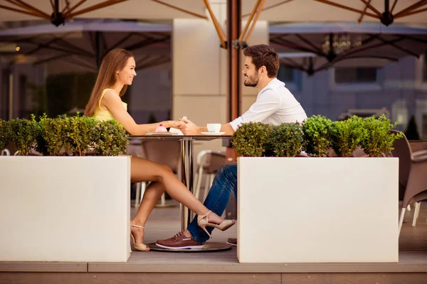 Playful girlfriend is flirting with guy by her leg, lovers are sitting in light cafe on a sunny day and enjoy coffee with tasies, talking, smiling, looking at each other — Stock Photo, Image