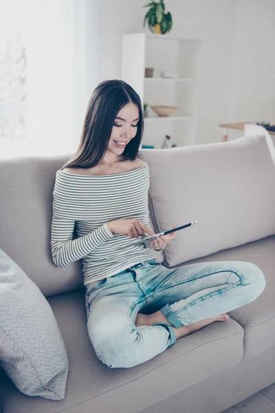 Närbild av beskurna fotot av unga glada kinesiska dam surfning på sin surfplatta, sitter på beige soffa inomhus i hemmet, klädd i casual outfit, med korsade ben — Stockfoto