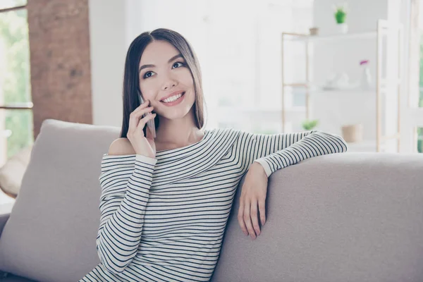 Upphetsad vacker Asiatisk tonåring med strålande leende pratar på hennes pda hemma. Hon bär casual kläder, sitta på mysiga beige soffa — Stockfoto