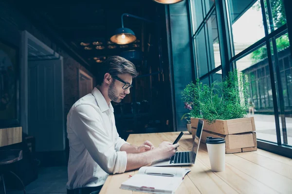 Achteraanzicht van het koelen van jonge hipster luisteren naar de muziek op zijn vakantie in grote witte moderne oortelefoons, in casual jeans slijtage, zitten in een donkere moderne Cafe in de buurt van het venster — Stockfoto