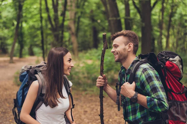 Cute excited lovers are hiking in a spring forest, talking and enjoying, wearing comfortable outfits for hiking, with backpacks and a stick — Stock Photo, Image