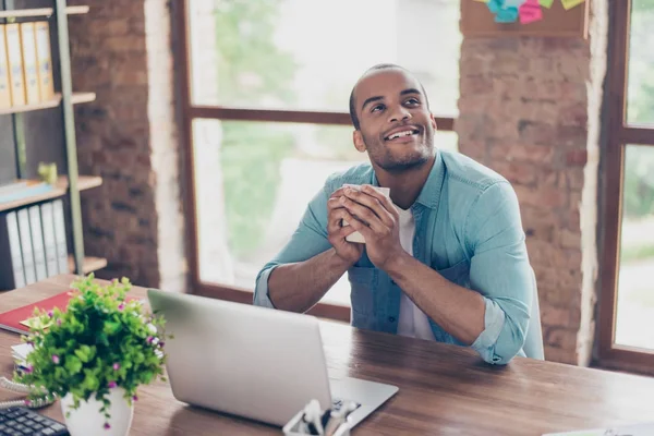 Jonge dromen mulat Amerikaanse arbeider denkt voor laptop op het werk plaats. Hij is gelukkig, glimlachen, achter hem is venster en bakstenen muur — Stockfoto