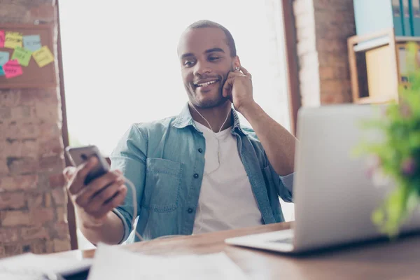 Portret van vrolijke jonge afro kerel die genieten van het luisteren naar mooie lied op zijn werkplek, op zijn slimme telefoon, dragen casual outfit, close-up glimlachen — Stockfoto