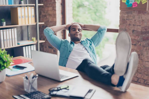 Unga glada afro freelancer vilar på en arbetsplats, med fötterna ovanpå skrivbordet, med slutna ögon, leende, drömmer — Stockfoto