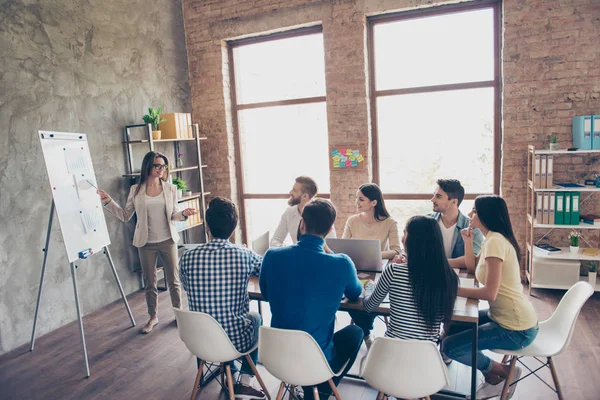 Young smart lady in glasses is reporting to the team of colleagues about the new project at the meeting with the white board. Workers are listening to her, all dressed in casual outfits — Stock Photo, Image