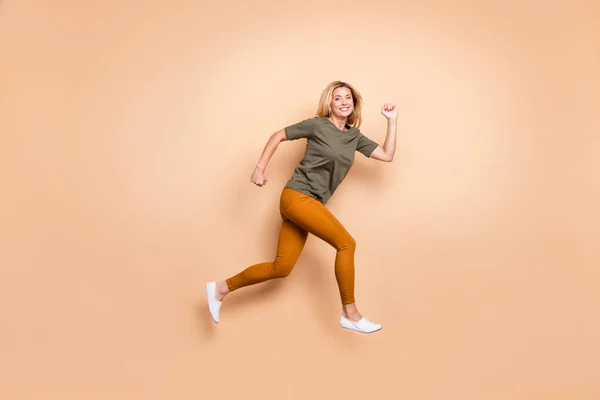 Ganzkörper-Profil-Foto von verrückter Dame springen Hochgeschwindigkeitsrennen Marathon Teilnehmer Wettkampfstimmung eilen ins Ziel tragen grünes T-Shirt gelbe Hose isoliert beige Farbhintergrund — Stockfoto