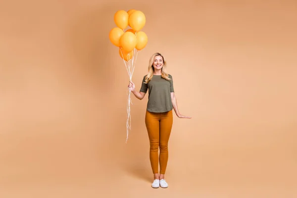 Full längd foto av fantastiska charmiga blond dam som håller många ljusa orange luft ballonger kom till födelsedagsfest bära grön t-shirt byxor isolerade beige färg bakgrund — Stockfoto