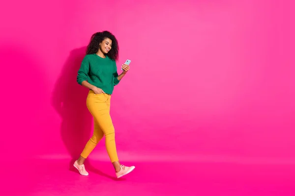 Full storlek profil sida foto av vågigt hår positiv glad flicka använda mobiltelefon gå promenad på hösten semester kyla bära grön pullover gula byxor sneakers isolerad rosa färg bakgrund — Stockfoto