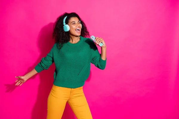 Retrato de pelo ondulado positivo afro chica americana tienen placer en las vacaciones de otoño escuchar canciones utilizar micrófono smartphone cantar usar pantalones amarillos jersey verde aislado color rosa fondo — Foto de Stock