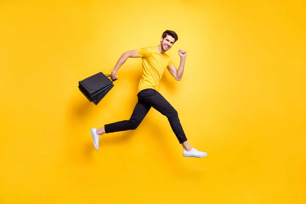 Foto em tamanho completo de cara bonito pulando alta segurando muitos pacotes velocidade apressando-se venda compras desgaste casual t-shirt calças pretas isolado cor amarela fundo — Fotografia de Stock