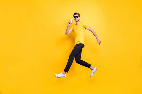 Full storlek foto av stilig kille hoppar högt innehav papper kopp takeaway kaffe promenader ner gatan bära sol specs casual t-shirt byxor isolerad gul färg bakgrund — Stockfoto