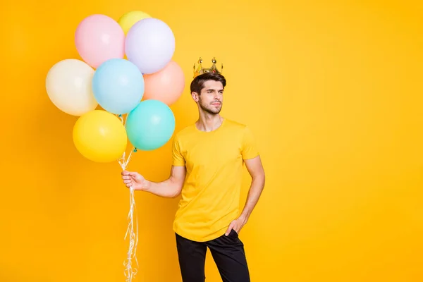 Foto de cara bonito gabar segurando balões de ar coroa dourada na cabeça mostra alto status social desgaste casual t-shirt calças pretas isolado cor amarela fundo — Fotografia de Stock