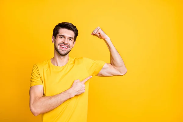 Foto de jovem bonito cara segurando punho levantou demonstrando perfeito grande bíceps indicando dedo no desgaste muscular casual t-shirt isolado cor amarela fundo — Fotografia de Stock