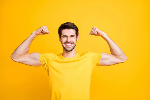 Photo of young handsome guy holding fists raised demonstrating perfect big biceps result after new sport gym wear casual t-shirt isolated yellow color background — ストック写真