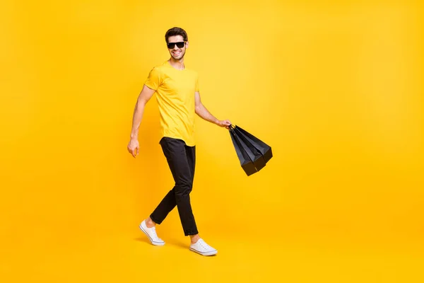 Full body foto van knappe man dragen boutique tassen maken in het buitenland winkelen gaan modieus winkelcentrum dragen casual t-shirt zwarte broek geïsoleerde gele kleur achtergrond — Stockfoto