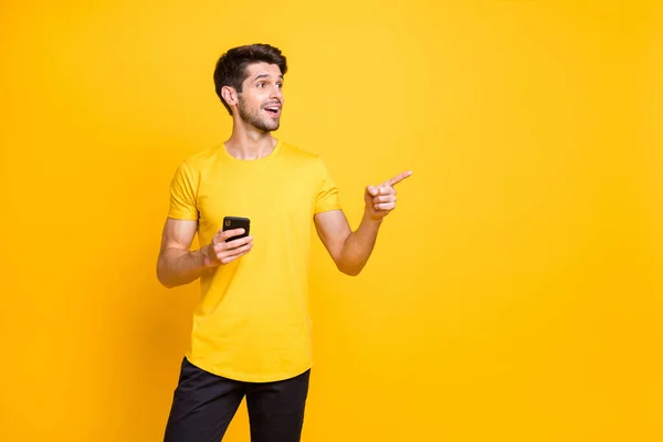 Photo of cheerful attractive handsome man wearing black trousers pointing at empty space with amazed facial expression holding telephone with hand isolated over vivid color background — ストック写真