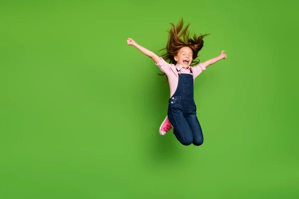 Full length photo of cheerful pretty little schoolchild jumping high rejoicing summer holidays hair flying wear casual denim overall pink shirt isolated green background — Stock Photo, Image