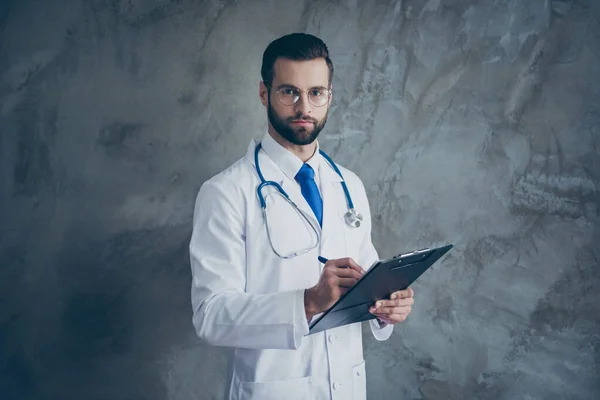 Porträt von konzentrierten Arzt Kerl halten sein Klemmbrett schreiben Patienten Beschwerden tragen weißen medizinischen Mantel isoliert über graue Farbe Hintergrund — Stockfoto