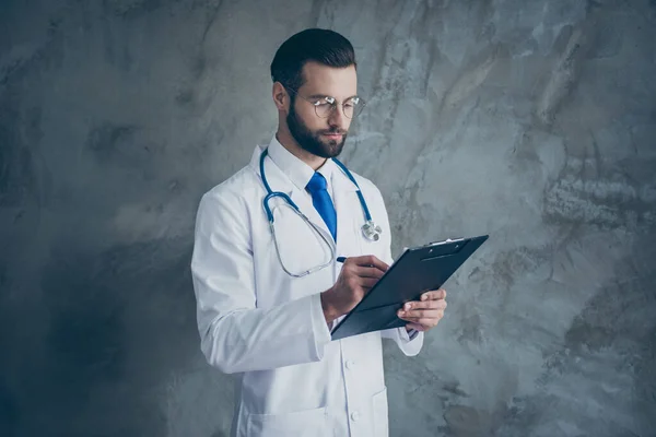 Profilseite Foto des fokussierten Chirurgen Mann hören Patienten Beschwerden schreiben in seinem Klemmbrett tragen weißen Arztkittel isoliert über grauem Hintergrund — Stockfoto