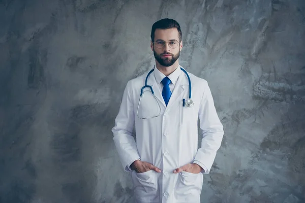 Retrato de médico talentoso grave colocar as mãos em bolsos de casaco branco olhar pronto para curar os pacientes na clínica isolada sobre fundo de cor cinza — Fotografia de Stock