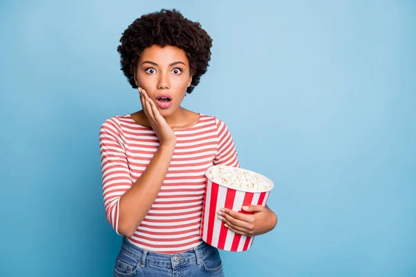 Photo of frightened feared girlfriend with popcorn in striped shirt jeans denim astonished while watching horror movie television curly wavy brown haired isolated pastel blue color background — Stock Photo, Image