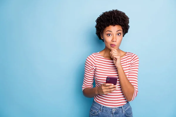 Photo of interested wondered pensive pondering girl browsing through telephone unsure about sending sms in striped shirt isolated pastel blue color background — Stock Photo, Image