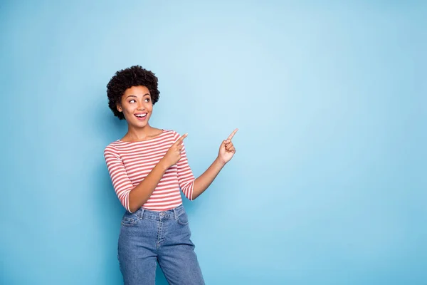 Photo of curly wavy cheerful positive nice pretty cute girlfriend pointing into empty space smiling toothily in jeans denim striped t-shirt isolated over blue pastel color background — ストック写真