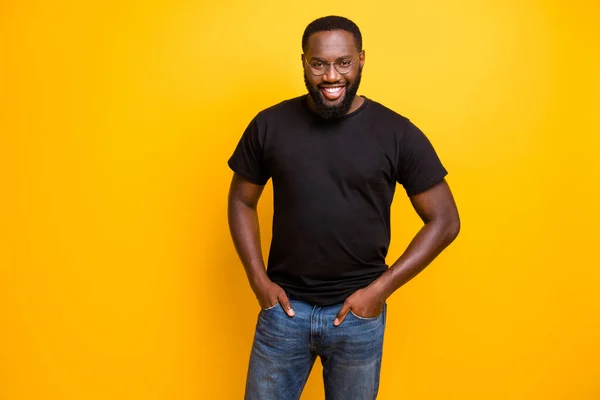 Foto de homem radiante de dentes alegre na moda com as mãos em jeans vestindo óculos de t-shirt isolados com as mãos em bolsos sobre fundo de cor vívida amarelo — Fotografia de Stock