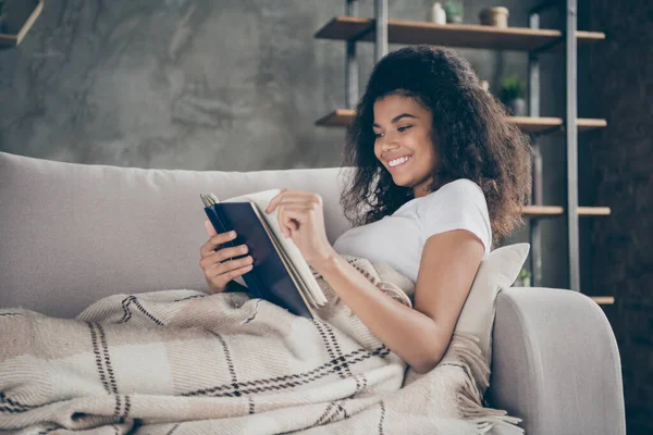 Profilo foto di pelle scura piuttosto sorprendente ondulato signora tenere libro lettura preferito storia d'amore romanzo storico sdraiato comodo divano coperto con plaid coperta soggiorno al coperto — Foto Stock