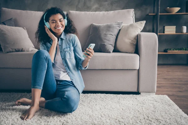 Foto de hermosa piel oscura dama ondulada ojos cerrados sosteniendo el uso de teléfono auriculares frescos escuchar nuevo audio sentado piso cerca de sofá ropa de mezclilla casual sala de estar en el interior — Foto de Stock