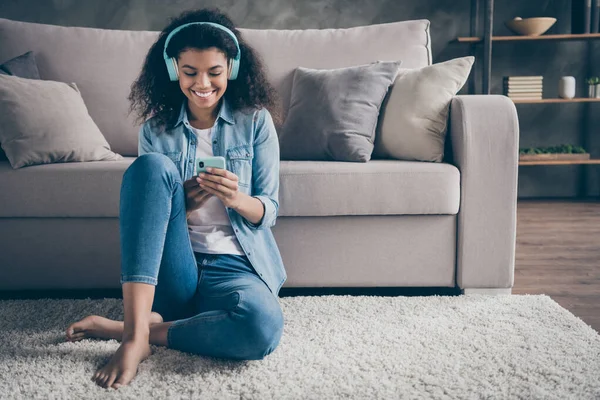 Foto di bella pelle scura ondulato signora homey umore tenere telefono usura fresco auricolari ascolto nuovo audio seduta piano vicino divano casual denim vestito soggiorno all'interno — Foto Stock