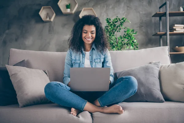 Fotografie docela ohromený tmavé kůže kudrnaté Lady notebook na kolena zkřížené nohy textové přátelé číst dobré zprávy sedí pohodlné gauč ležérní džínové oblečení obývací pokoj uvnitř — Stock fotografie