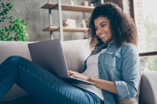 Closeup profile photo of pretty amazed dark skin curly lady notebook on knees texting friends read cool good news lying comfy couch casual denim outfit living room indoors — ストック写真