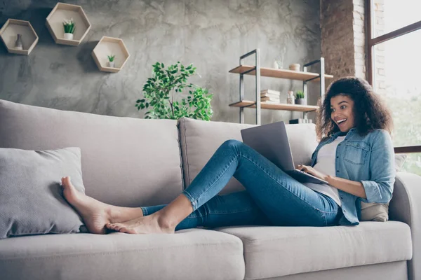 Foto de perfil de la señora rizada piel oscura bastante sorprendido mantener mensajes de texto portátil con amigos leer buenas noticias frescas tumbado cómodo sofá ropa de mezclilla casual sala de estar en el interior —  Fotos de Stock