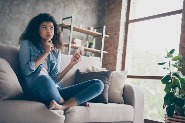 Photo of pretty dark skin curly lady hold telephone deeply minded think over new port text finger on chin sitting comfy couch casual denim outfit living room indoors — Stock Photo, Image