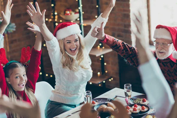 Foto de alegre mujer bonita positiva con hija cercana con el abuelo en la cabeza del sombrero de santa en la mesa con la comida festiva y la iluminación de luces detrás — Foto de Stock