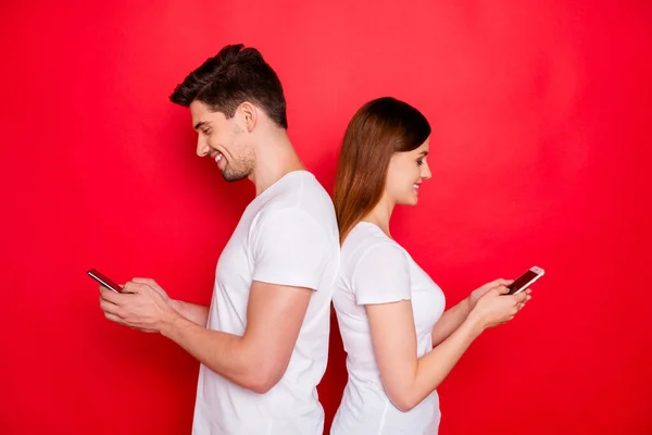 Foto av flickvän pojkvän står tillbaka till ryggen chatta med varandra bläddra genom telefonen ler tandlöst bär vit t-shirt isolerad levande färg bakgrund — Stockfoto