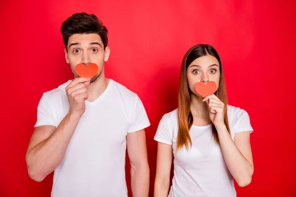 Foto de alegre positivo agradável muito bonito casal de pessoas cobrindo sua boca com pequenos corações vermelhos de amor vestindo t-shirt branca isolado fundo de cor vívida — Fotografia de Stock