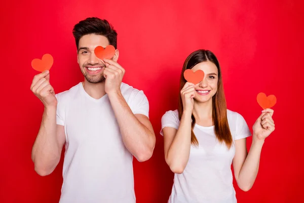 Foto de alegre positiva bonita pareja bonita de novio novia sonriente cubriendo su ojo mostrando su amor con corazón rojo blanco postales camiseta aislado color vivo fondo —  Fotos de Stock