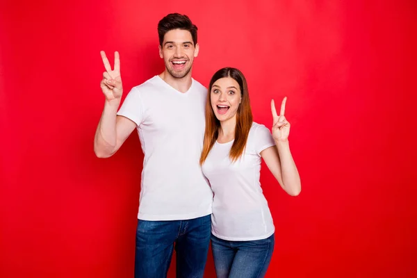 Foto de lindo novio bonita pareja que le muestra dos signos de v sonriendo toothily pelo rojo morena en jeans vaqueros camiseta blanca aislada sobre fondo de color brillante —  Fotos de Stock