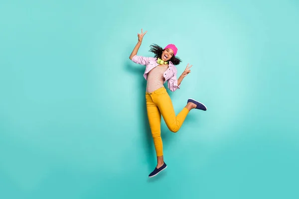 Full length body size photo of casual cute nice girl jumping pokazujące podwójne v-sign żółte spodnie czapka nakrycie głowy różowy marynarka paski t-shirt podekscytowany izolowany żywy kolor ciemny tło — Zdjęcie stockowe