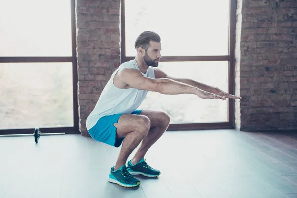 Foto de perfil de cuerpo completo de macho chico haciendo sentadillas estáticas proceso de quema de grasa ropa deportiva tanque-top shorts zapatillas casa de entrenamiento estudio cerca de grandes ventanas en el interior — Foto de Stock