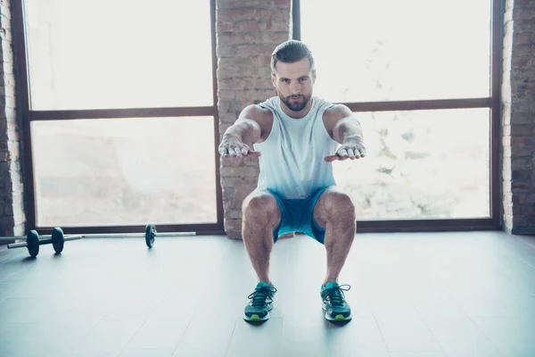 Foto de cuerpo entero de macho chico haciendo sentadillas estáticas quemar grasa ropa deportiva blanco tanque-top azul pantalones cortos zapatillas de deporte de entrenamiento en casa estudio cerca de grandes ventanas en el interior —  Fotos de Stock