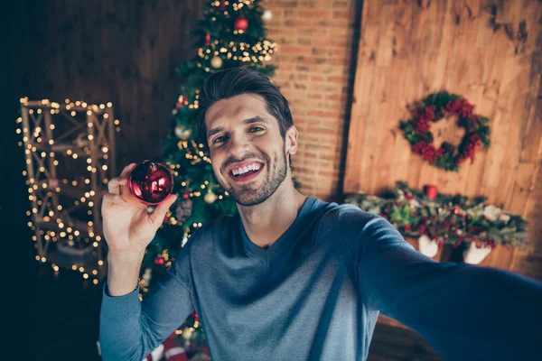 Auto foto de alegre engraçado animado homem segurando Natal brinquedo bugiganga com as mãos sorrindo toothily tomar selfie para promover a bola na frente da árvore — Fotografia de Stock