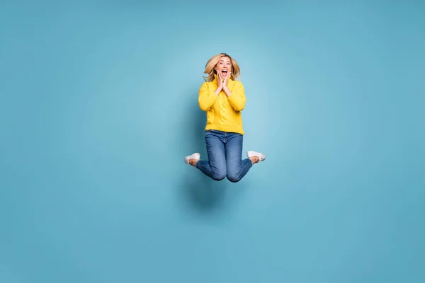 Corpo inteiro foto de senhora louca pulando alto celebrando viagem livre no exterior regozijando as mãos em maçãs do rosto desgaste malha amarelo pulôver jeans isolado azul cor fundo — Fotografia de Stock