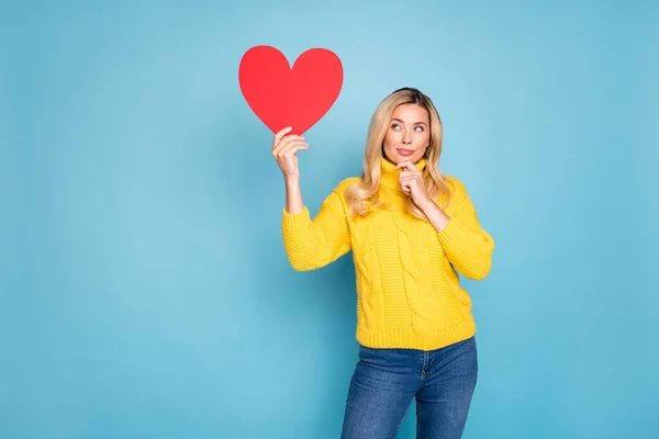 Foto de la señora rubia increíble sosteniendo el corazón de papel rojo grande pensando sobre la respuesta correcta en la fecha invitación desgaste de punto pantalones vaqueros de jersey amarillo aislado fondo de color azul —  Fotos de Stock