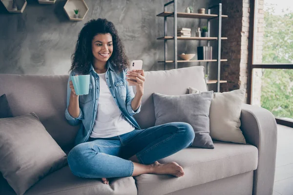 Foto de hermosa piel oscura dama ondulada ambiente hogareño celebración de la taza de bebida caliente teléfono freelance red social sentado acogedor sofá ropa de mezclilla casual sala de estar en el interior — Foto de Stock