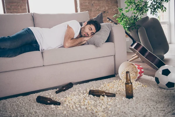 Photo of multiethnic boozer alcoholic guy lying sofa sleeping hangover beer empty bottles popcorn floor suffering after bachelor party morning messy rubbish dirty flat indoors — Stock Fotó