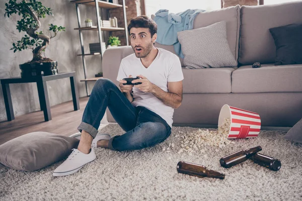Foto de tipo árabe de raza mixta sentado alfombra sofá inclinado mantenga joystick jugando videojuego emocionado quiere ganar botellas de cerveza vacías palomitas de maíz en el suelo desorden desordenado plana en interiores — Foto de Stock